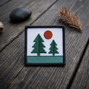 May include: A square embroidered patch featuring a white background with two dark green pine trees and a solid burnt orange circle representing the sun. The bottom section is a solid teal green band, and the entire patch is bordered in black.