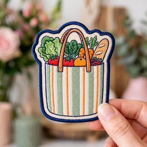 May include: Embroidered patch featuring a shopping bag filled with groceries. The bag has green and orange stripes, a brown handle, and is overflowing with lettuce, tomatoes, carrots, an orange, bread, and berries. The patch has a blue border.