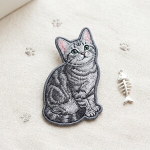 May include: A gray tabby cat embroidered patch featuring detailed stitching, green eyes, and pink inner ears, accompanied by a small silver-toned metal fish skeleton charm. The cat is depicted in a seated position with a striped tail curled around.