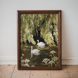 May include: A framed painting of two white swans swimming in a pond surrounded by green foliage. The swans are in the center of the painting, with the foliage surrounding them. The painting is in a dark brown frame.