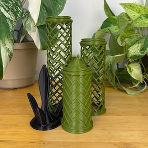 Op de afbeelding: Drie groene plastic plantenondersteuningen, twee zijn cilindrisch met een traliewerkontwerp en één is een massieve cilinder met een chevron-patroon. Een zwarte plastic plantenondersteuning bevindt zich op de voorgrond.