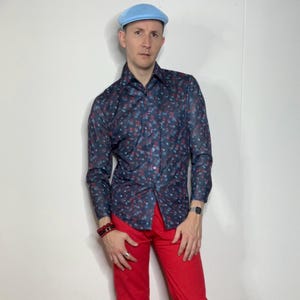 May include: A man wearing a blue patterned long-sleeve shirt with a light blue newsboy cap and red pants.