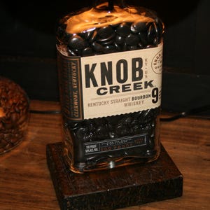 May include: A bottle of Knob Creek Kentucky Straight Bourbon Whiskey, 9 years old, on a dark wooden stand. The label is cream-colored with black text. The bottle is filled with dark, round objects.