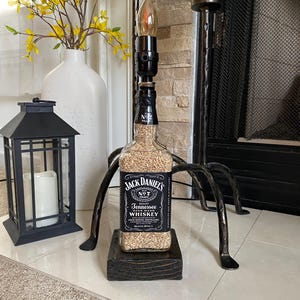 May include: A repurposed Jack Daniel's whiskey bottle lamp with a black metal base and a clear glass shade. The bottle is filled with small, white grains and has the Jack Daniel's label on it. The lamp is sitting on a wooden base.