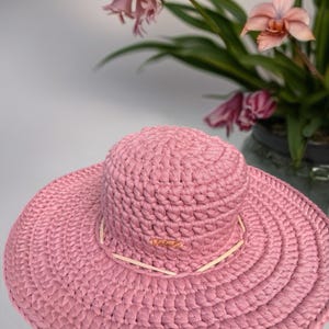May include: A wide-brimmed, pink sun hat with a woven texture. The hat features a light-colored band around the crown and a small, gold-toned logo. The hat is displayed in front of a potted orchid with pink flowers.