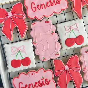 Puede incluir: Surtido de galletas de azúcar decoradas sobre una rejilla de enfriamiento. Las galletas incluyen formas de lazo rosa, galletas con el número 5 y galletas con el nombre "Genesis". Otras galletas presentan cerezas.