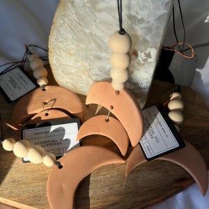 May include: Four terracotta crescent moon essential oil diffusers with wooden beads and black string. Each diffuser has a label that reads "Mindful Made Co. Essential Oil Diffuser".
