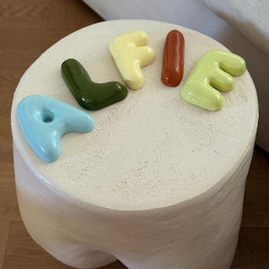 May include: A cream-coloured, rounded stool with a textured surface. Ceramic letters in various colours spell "LIFE" on top: light blue "L", dark green "I", yellow "F", and orange "E". The stool has a unique, organic shape.