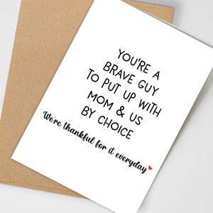 May include: A white greeting card with black text that reads "YOU'RE A BRAVE GUY TO PUT UP WITH MOM & US BY CHOICE. We're thankful for it everyday" is placed on a brown envelope. A small red heart is at the end of the text.