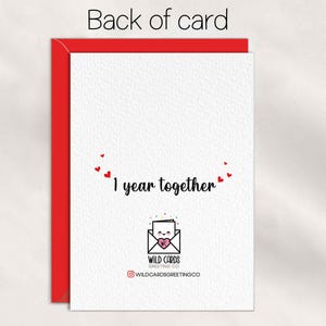 May include: A white greeting card with a red envelope behind it. The card features the text 1 year together in black script, surrounded by small red hearts. Below, a logo shows an envelope with a face and a pink heart, labeled WILD CARDS GREETING CO.