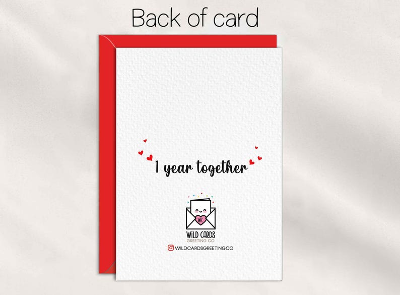 May include: A white greeting card with a red envelope behind it. The card features the text 1 year together in black script, surrounded by small red hearts. Below, a logo shows an envelope with a face and a pink heart, labeled WILD CARDS GREETING CO.
