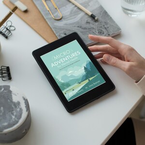 May include: A black e-reader showing the cover of "Micro Adventures for Mental Health", featuring a landscape illustration. The e-reader is on a white surface, with stationery and a hand nearby.