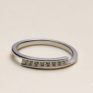 May include: A silver ring with a curved design. The ring has a row of small, clear gemstones set along the curved section. The ring is displayed on a light-coloured surface.