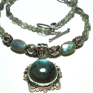 May include: A silver necklace with a large oval labradorite pendant. The necklace is made with oval labradorite beads and silver spacers. The pendant is set in a silver frame with a detailed design.