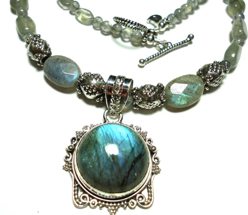 May include: A silver necklace with a large, round, blue labradorite gemstone pendant. The necklace is made with silver beads and labradorite stones.
