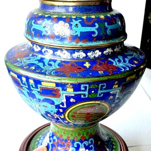 May include: A blue and gold cloisonn&eacute; vase with a floral and geometric design. The vase is on a wooden stand with intricate carvings.