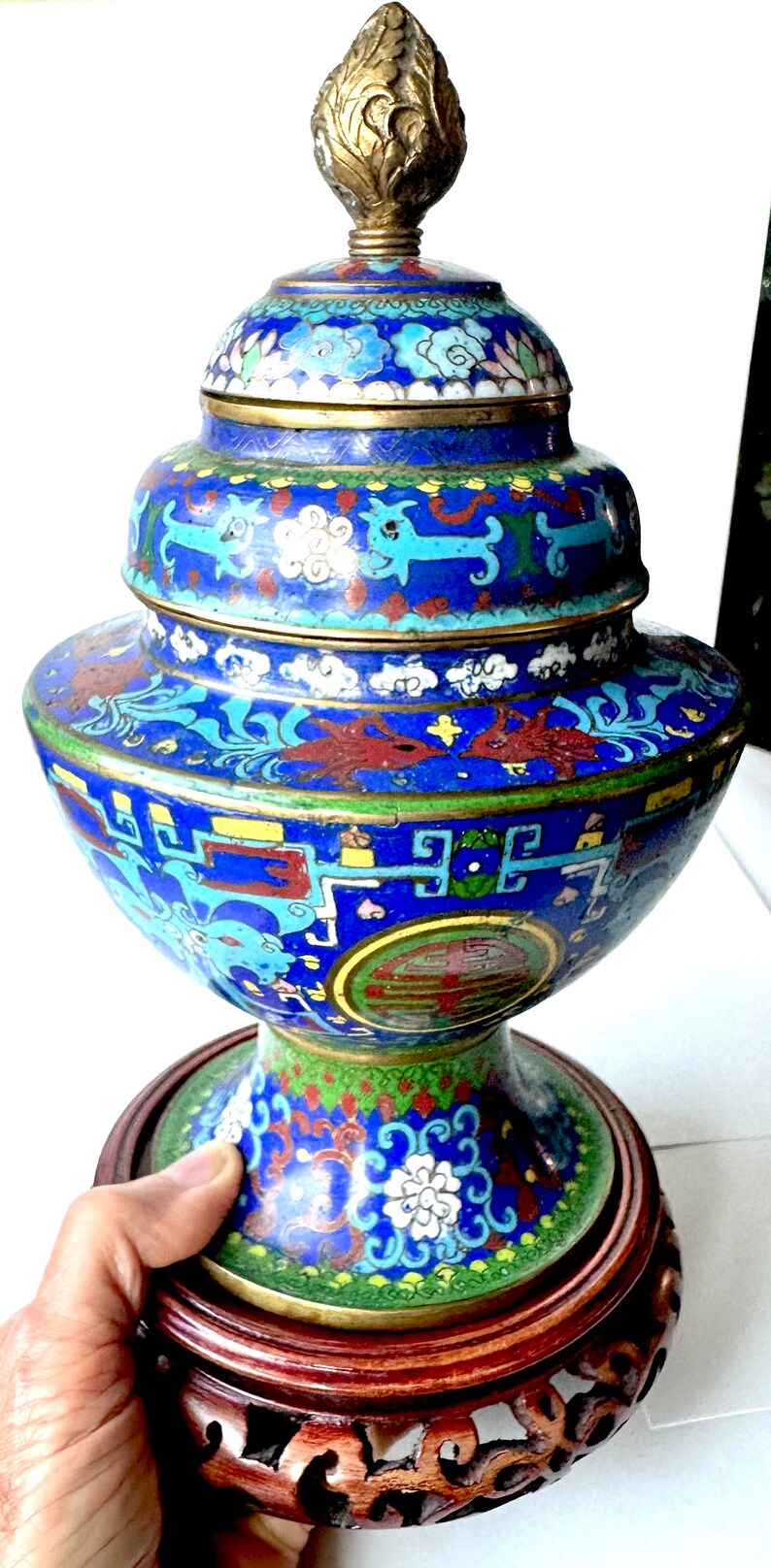 May include: A blue and gold cloisonn&eacute; vase with a floral and geometric design. The vase is on a wooden stand with intricate carvings.