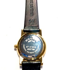 May include: A gold-tone Movado watch with a black leather band. The watch face is round and has a silver dial. The back of the watch has the Movado logo and the words "Swiss Made" and "87 A1845 8307768".