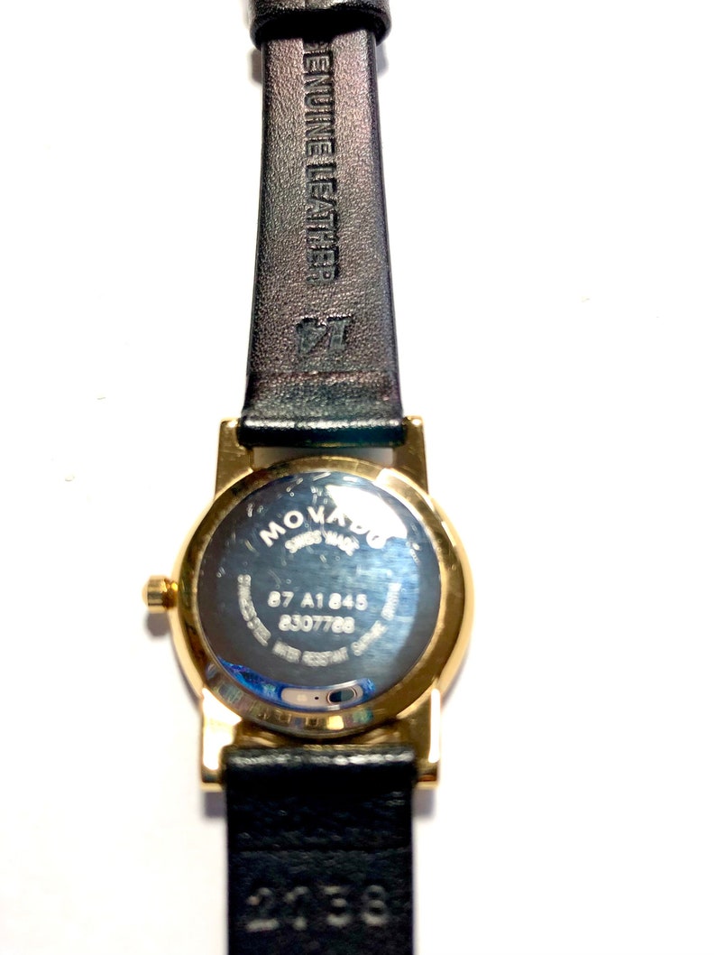 May include: A gold-tone Movado watch with a black leather band. The watch face is round and has a silver dial. The back of the watch has the Movado logo and the words "Swiss Made" and "87 A1845 8307768".