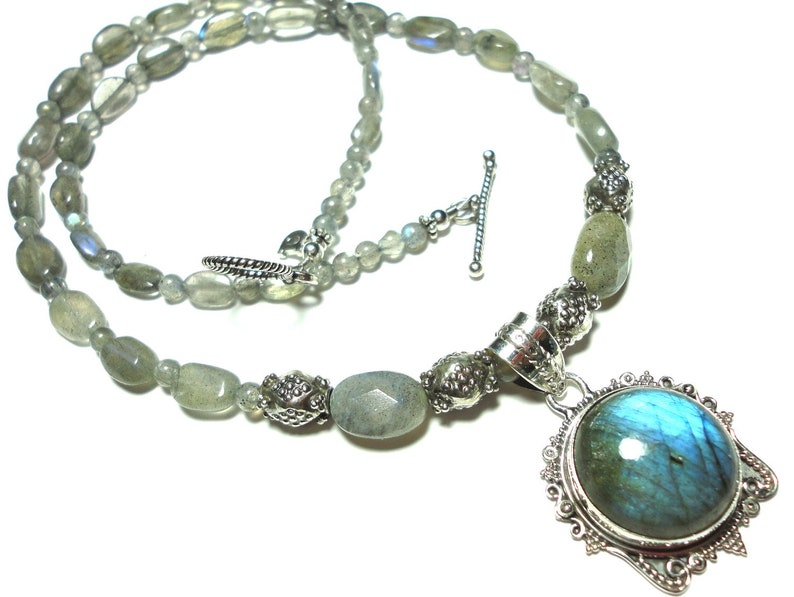 May include: A silver necklace with a large, round, iridescent blue labradorite gemstone pendant. The necklace is made with oval-shaped, light gray labradorite beads and silver accents.