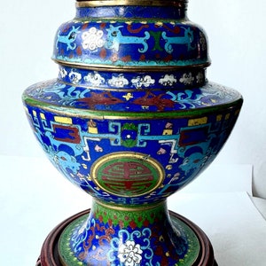 May include: A blue and gold cloisonn&eacute; vase with intricate floral and geometric designs. The vase is on a wooden stand with carved details.