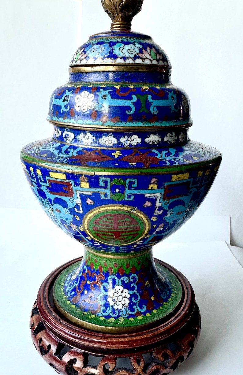 May include: A blue and gold cloisonn&eacute; vase with intricate floral and geometric designs. The vase is on a wooden stand with carved details.