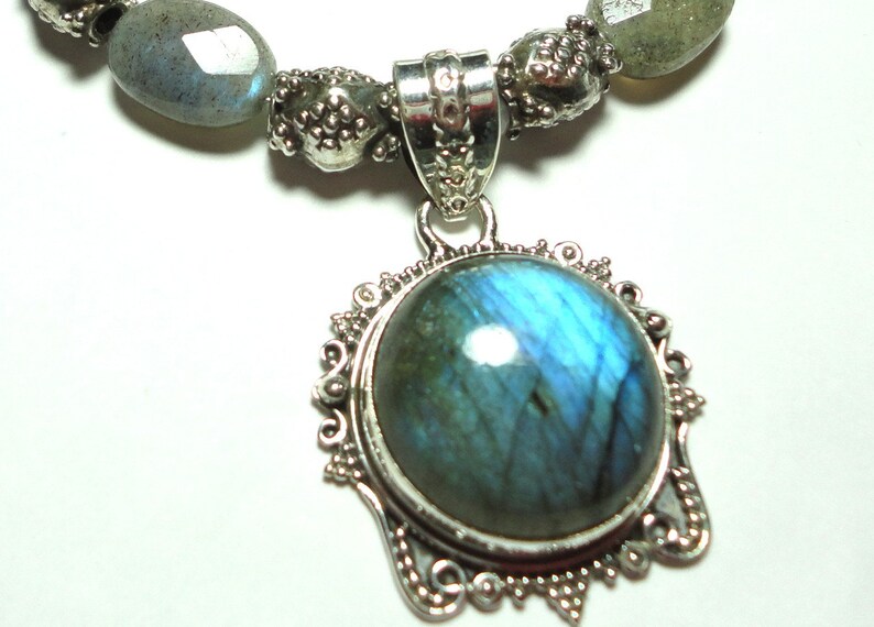 May include: A silver necklace with a large, round, blue labradorite gemstone pendant. The pendant is set in a silver filigree frame. The necklace is made of silver beads and labradorite stones.
