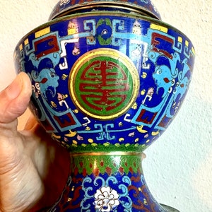 May include: A blue and gold cloisonn&eacute; incense burner with a lid. The burner is decorated with intricate floral and geometric patterns, and features a large, circular symbol in the center. The burner is on a pedestal with a similar design.