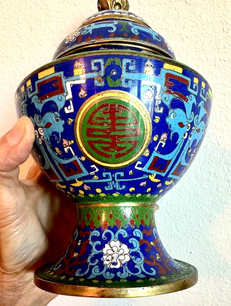 May include: A blue and gold cloisonn&eacute; incense burner with a lid. The burner is decorated with intricate floral and geometric patterns, and features a large, circular symbol in the center. The burner is on a pedestal with a similar design.