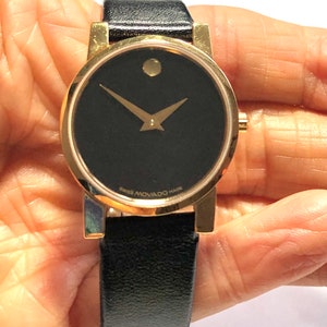 May include: A gold-tone Movado watch with a black face and black leather band. The watch face features a single dot at the 12 o'clock position and gold-tone hands. The watch band is a simple black leather strap.