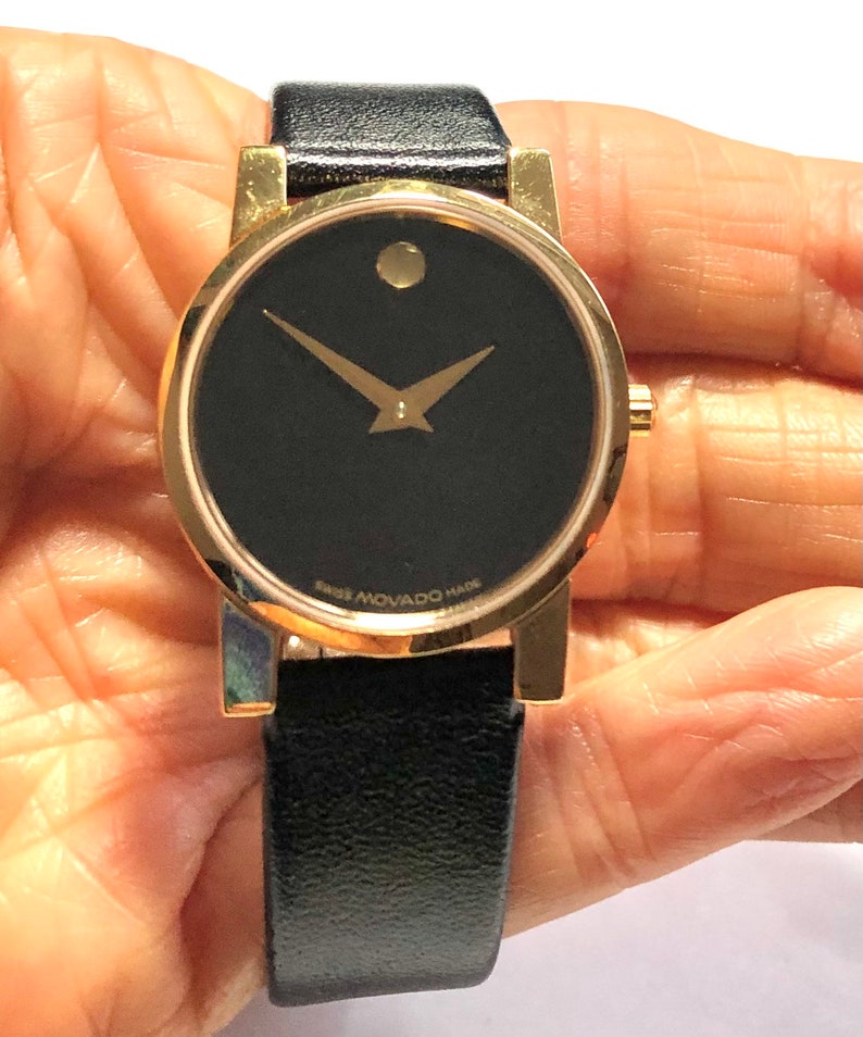 May include: A gold-tone Movado watch with a black face and black leather band. The watch face features a single dot at the 12 o'clock position and gold-tone hands. The watch band is a simple black leather strap.
