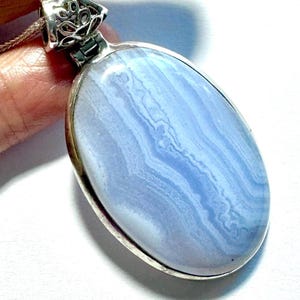 May include: An oval-shaped blue lace agate pendant set in a polished silver frame. The gemstone features light blue and white banded patterns throughout its surface. The pendant includes a decorative silver bail with an intricate cutout design.