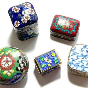 Może przedstawiać: Pięć małych ozdobnych pudełek z misternymi wzorami kwiatowymi i geometrycznymi. Pudełka są wykonane z metalu i mają wykończenie emaliowane cloisonn&eacute;. Kolory obejmują niebieski, czerwony, zielony, biały i czarny.