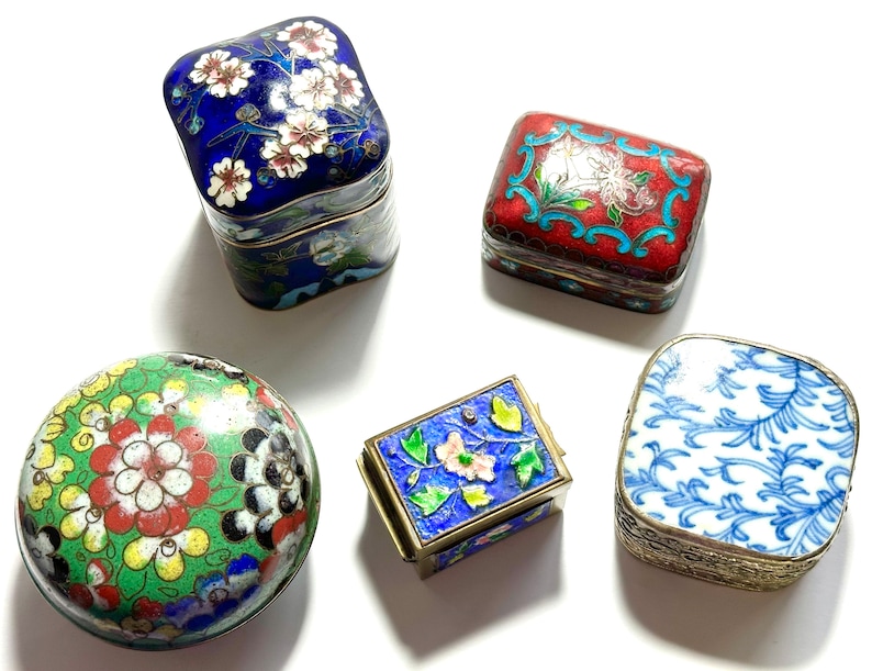 Może przedstawiać: Pięć małych ozdobnych pudełek z misternymi wzorami kwiatowymi i geometrycznymi. Pudełka są wykonane z metalu i mają wykończenie emaliowane cloisonn&eacute;. Kolory obejmują niebieski, czerwony, zielony, biały i czarny.