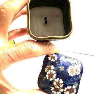 Może przedstawiać: Kwadratowe pudełeczko na drobiazgi wykonane w technice cloisonn&eacute;, z niebieską emaliowaną podstawą i kwiatowym wzorem w kolorze białym, r&oacute;żowym i złotym. Pokrywka jest otwarta, ukazując białą świeczkę w środku.