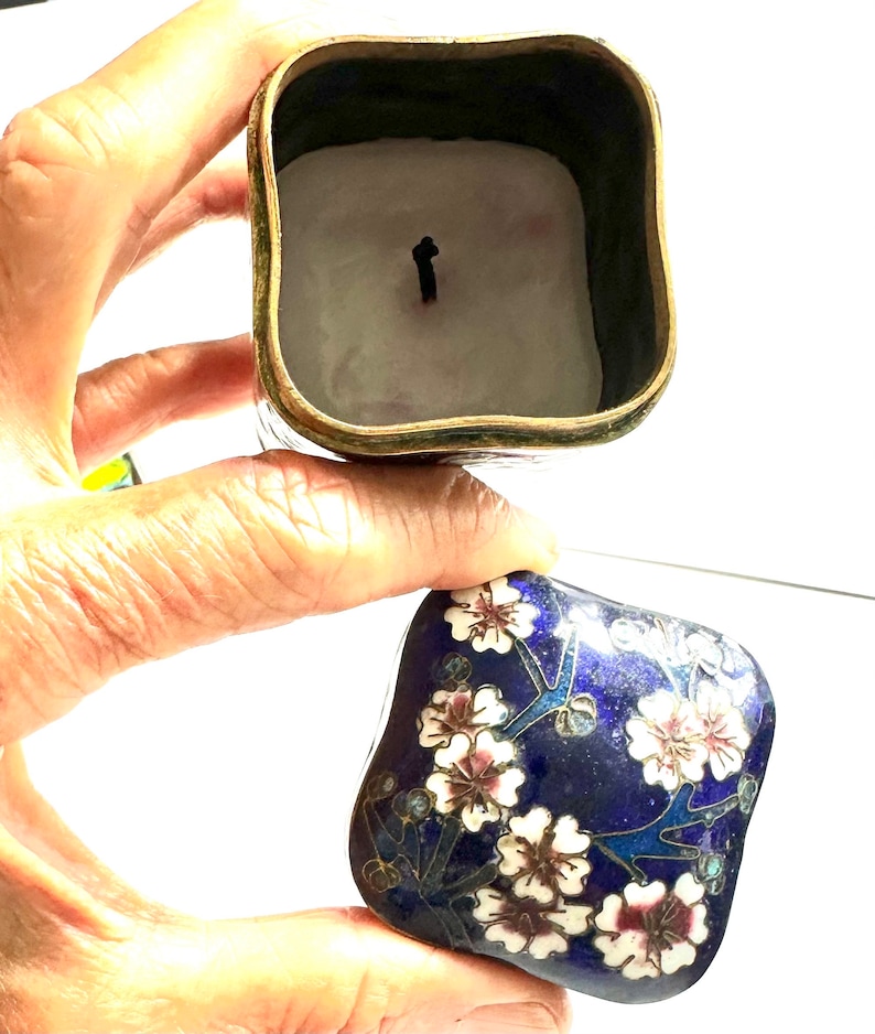 Może przedstawiać: Kwadratowe pudełeczko na drobiazgi wykonane w technice cloisonn&eacute;, z niebieską emaliowaną podstawą i kwiatowym wzorem w kolorze białym, r&oacute;żowym i złotym. Pokrywka jest otwarta, ukazując białą świeczkę w środku.