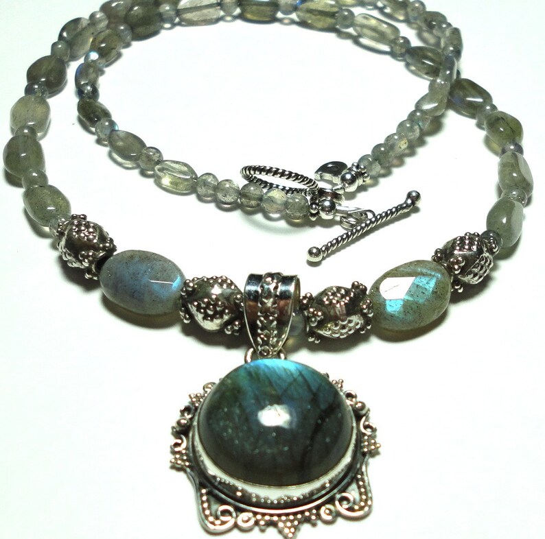 May include: A silver necklace with a large oval labradorite pendant. The necklace is made with oval labradorite beads and silver spacers. The pendant is set in a silver frame with a detailed design.