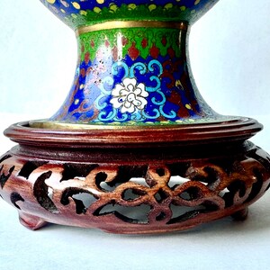 May include: A blue and green cloisonn&eacute; vase with intricate floral designs sits on a carved wooden stand. The vase has a wide mouth and a round body.