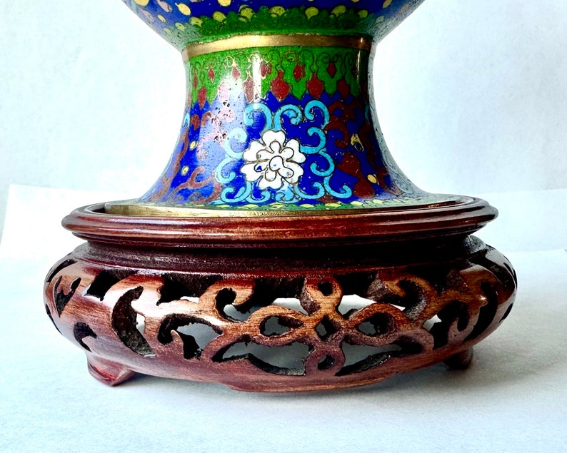 May include: A blue and green cloisonn&eacute; vase with intricate floral designs sits on a carved wooden stand. The vase has a wide mouth and a round body.