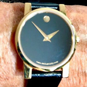 May include: A gold-tone wristwatch with a black face and black leather band. The watch has a simple design with a single hand and a small diamond-shaped marker at the 12 o'clock position. The watch face also has the text "Movado" printed on it.
