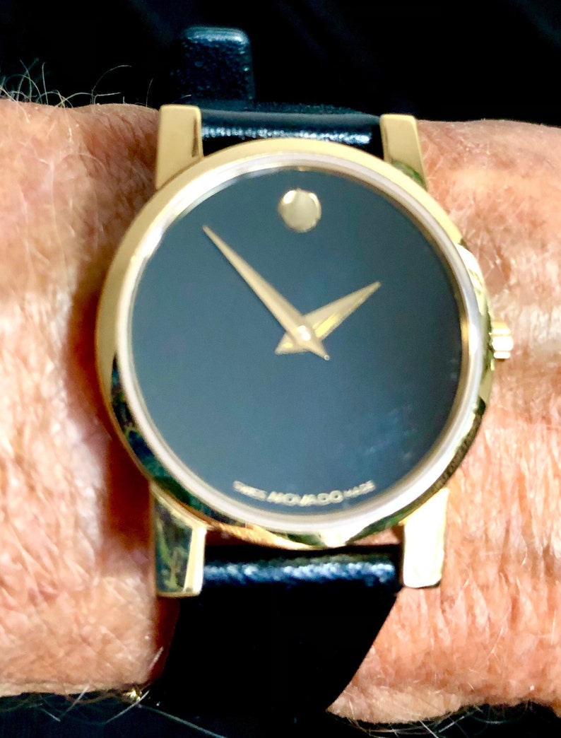 May include: A gold-tone wristwatch with a black face and black leather band. The watch has a simple design with a single hand and a small diamond-shaped marker at the 12 o'clock position. The watch face also has the text "Movado" printed on it.