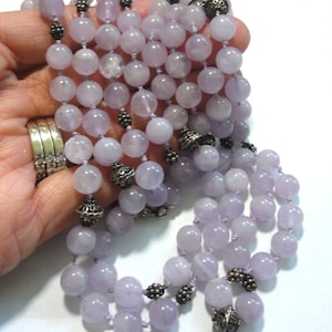 May include: A long lavender colored beaded necklace with silver accents. The necklace is made of round beads and has a tassel at the end.