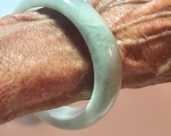 May include: A smooth, polished bangle bracelet crafted from pale green jade. The circular accessory features a translucent, milky appearance with subtle variations in color intensity throughout the stone, showcasing a rounded, thick profile.