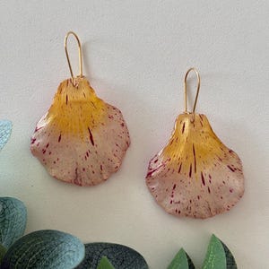May include: A pair of dangle earrings featuring petal-shaped components with a gradient of yellow at the top transitioning to a pale pink base, accented with scattered dark red speckles and attached to thin gold-toned metal ear wires.