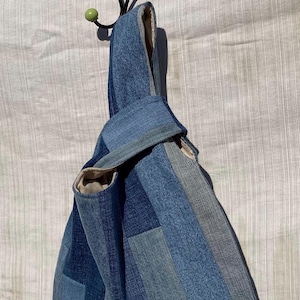 May include: A Japanese-style knot bag constructed from various shades of blue denim fabric. The bag features a patchwork design with vertical panels of light, medium, and dark blue denim, a beige interior lining, and a single handle formed by a knot.