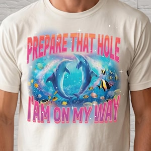 Può includere: T-shirt color avorio con una grafica raffigurante due delfini, pesci e la scritta rosa "PREPARE THAT HOLE I AM ON MY WAY". Il disegno è ambientato su uno sfondo blu e verde.