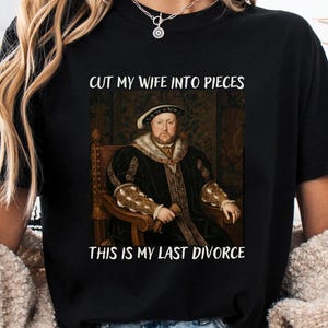 May include: Black t-shirt featuring a portrait of a historical figure with the text "CUT MY WIFE INTO PIECES" and "THIS IS MY LAST DIVORCE". The shirt has a round neckline and short sleeves.