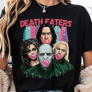 May include: Black t-shirt featuring a graphic of four characters blowing bubble gum, with the text "DEATH EATERS" in pink. The graphic has a colorful background. The shirt is a comfortable, casual style.