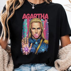 May include: Black t-shirt with the word "AGARTHA" in pink above a graphic of a person with long blonde hair blowing a bubblegum bubble. The person is wearing a blue and gold uniform and holding up a hand with a purple star.
