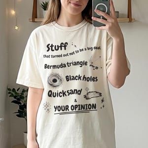 Può includere: Una t-shirt a maniche corte color avorio con testo nero che recita: Stuff that turned out not to be a big deal, Bermuda triangle, Black holes, Quicksand & YOUR OPINION, accompagnato da piccole illustrazioni grafiche nere di spazio e natura.